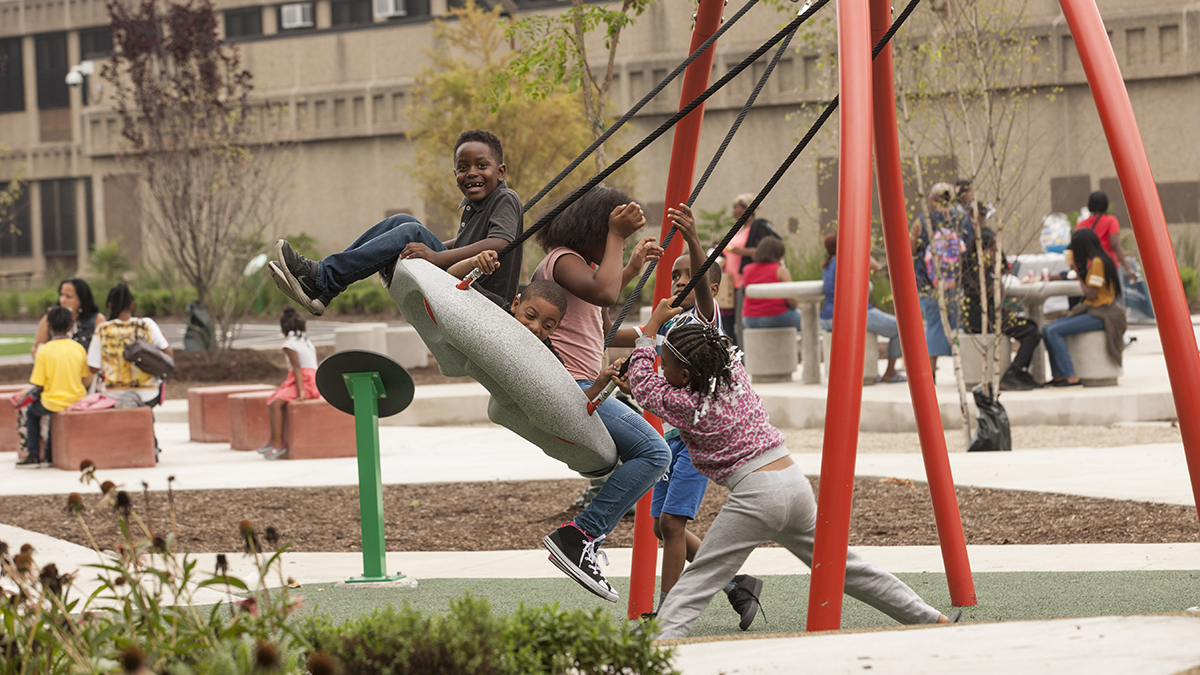 Children swing on a multi-person swing set at the John W. Cook Academy schoolyard, while adults sit in the background.