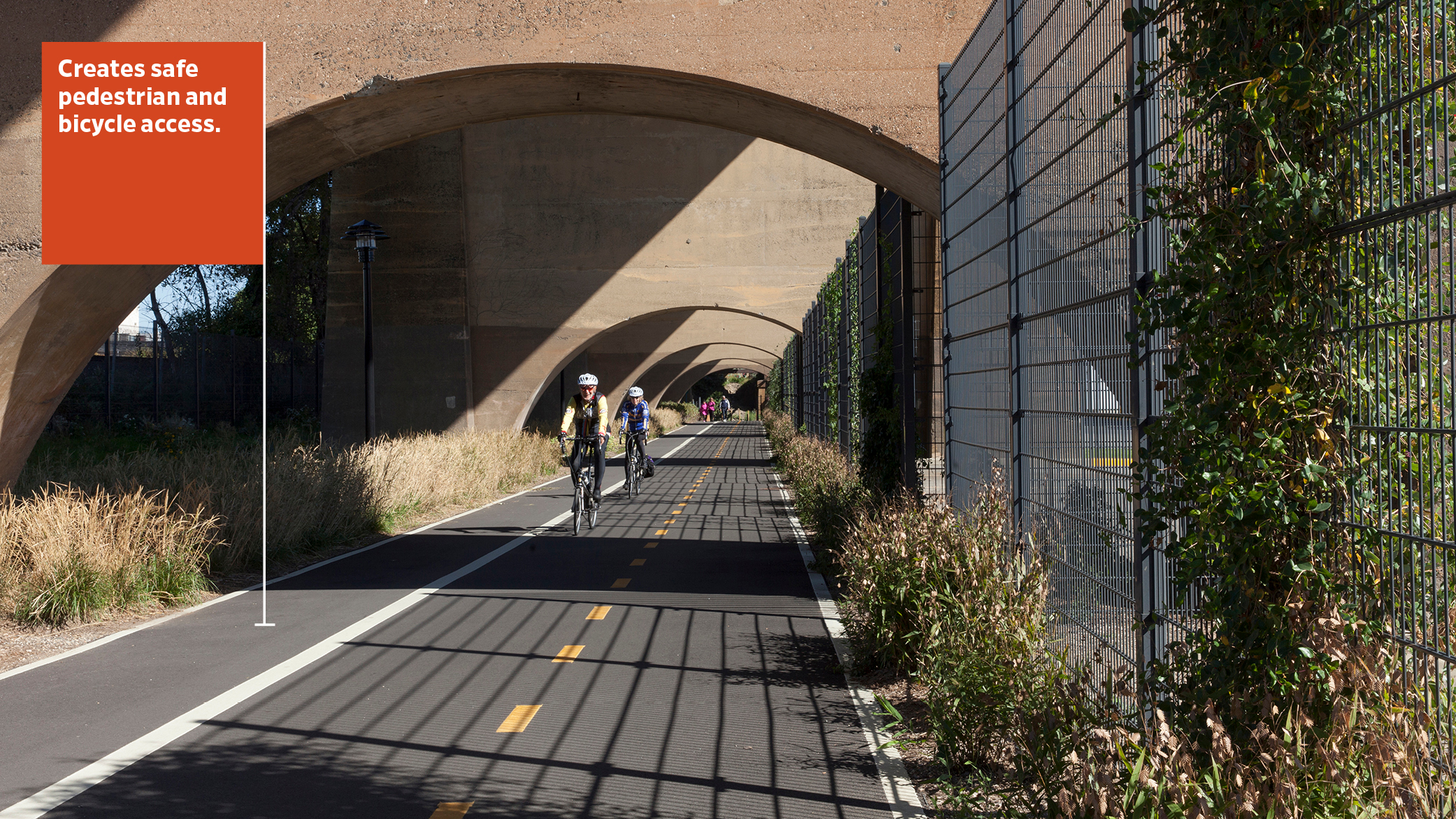 Two bicyclists enjoy riding on the pathway of the Randall’s Island Connector; at right, vines crawl up fence.